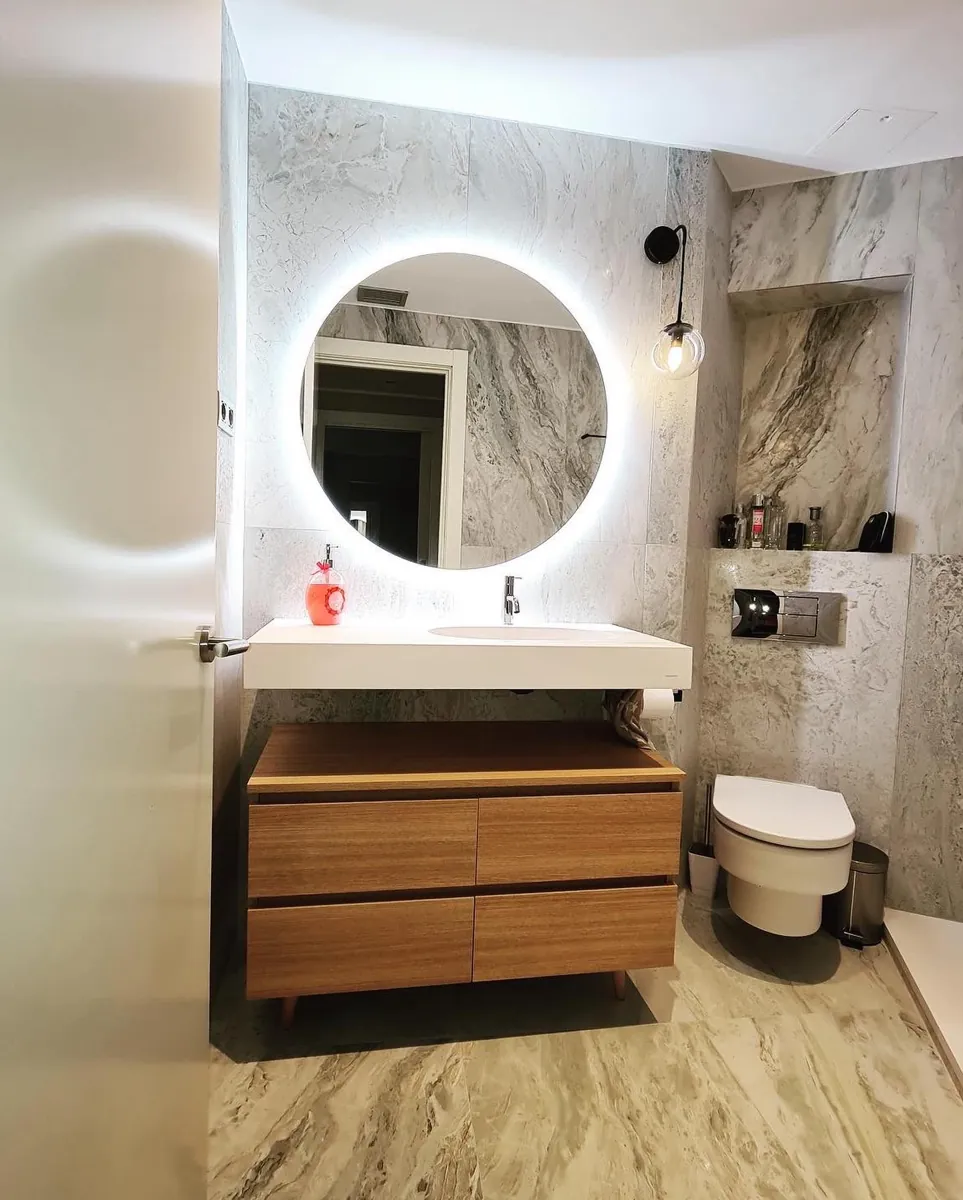 Baño con mueble de madera suspendido, espejo redondo retroiluminado y ducha de obra con mampara