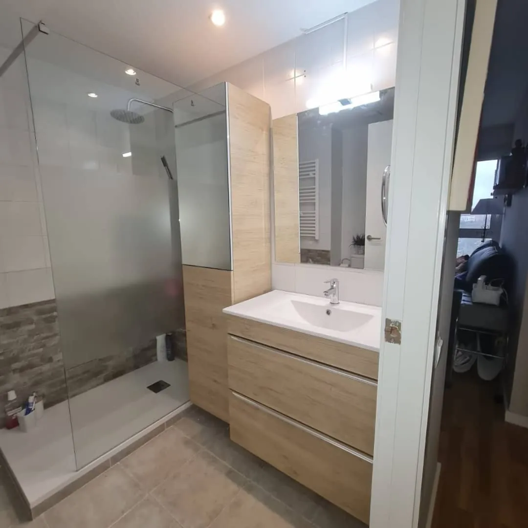 Ducha a ras de suelo con mampara de vidrio, mueble de madera clara y radiador toallero