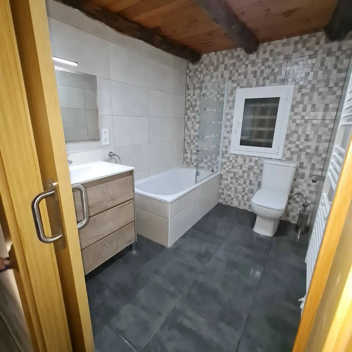 Después: Baño rehecho conservando las vigas de madera, con alicatado hasta el techo, mosaico, bañera nueva y radiador toallero