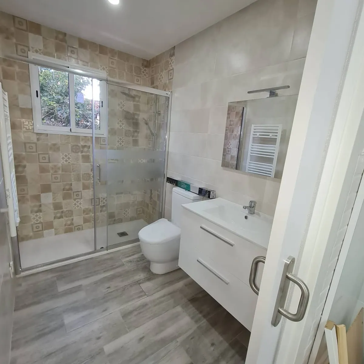 Después: Baño antiguo renovado por completo: ducha con mampara corredera, mueble suspendido blanco y suelo efecto madera