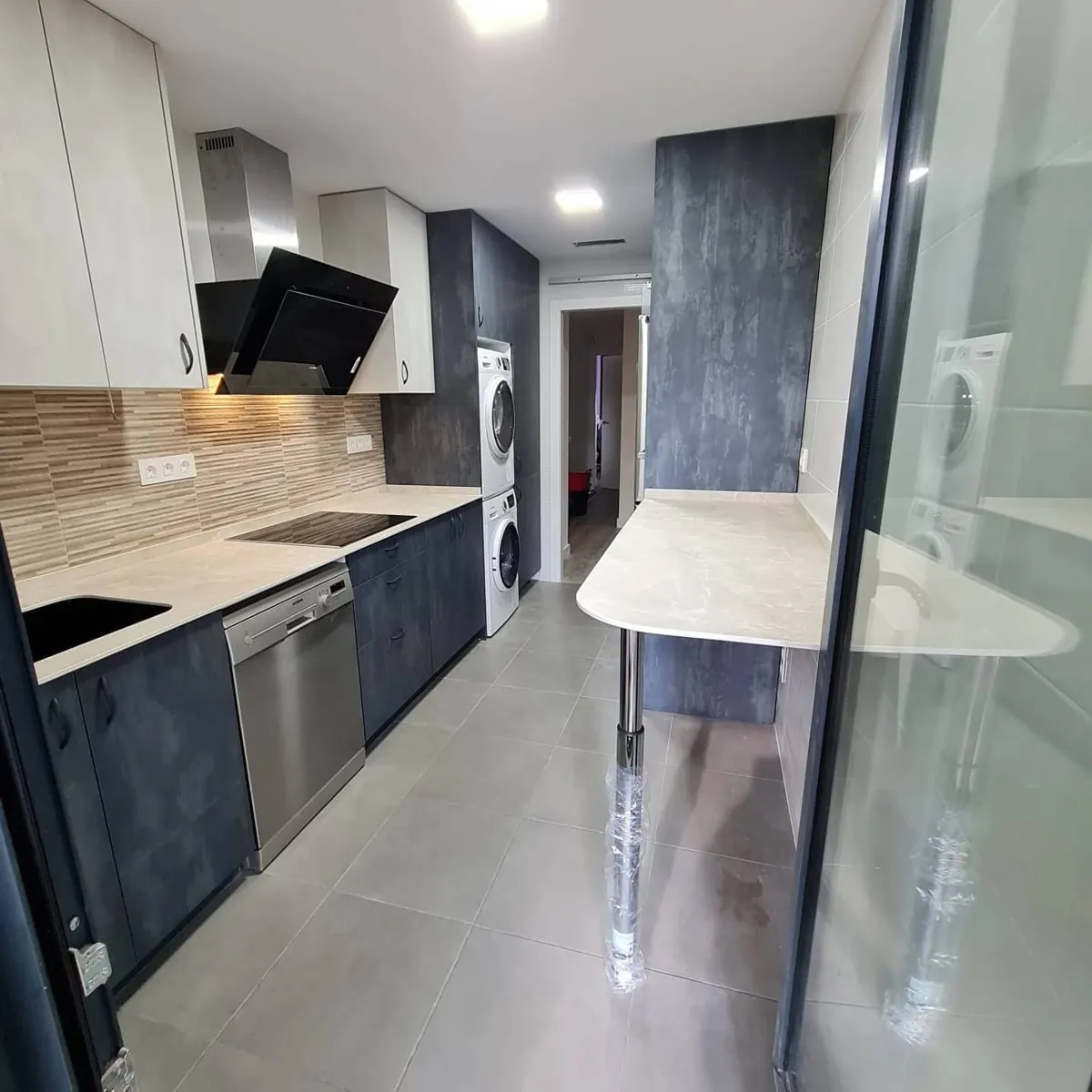 Cocina lineal con frentes grises y madera, campana negra y lavadora integrada